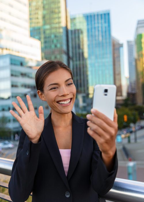 People on video call saying hello waving at camera. Asian woman video calling business partner on smartphone online in city street – Office buidlings on videochat call. Selfie entrepreneur cellphone. People on video call saying hello waving at camera. Asian woman video calling business partner on smartphone online in city street - Office buidlings on videochat call. Selfie entrepreneur cellphone.